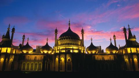 A building with several spires and domes is blackened against a purple sunset. 