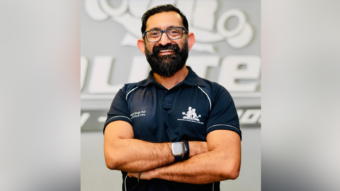 Health Scientist & Body Transformation Coach, Manjit Singh Dol, with dark hair, a beard and glasses smiling with his arms folded and wearing a black t-shirt against a faded grey background.