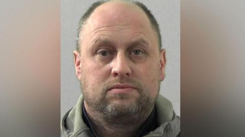 Mugshot of Stephen York. He is balding with very short dark hair, a thin wispy fair beard with grey hints and is wearing a grey hoodie.