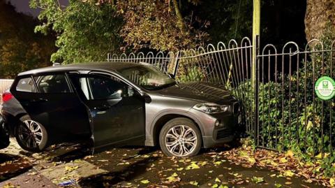 A BMW is sideways on and crashed into a fence. It is dark outside and there are leaves on the floor. The driver and back driver doors are open 