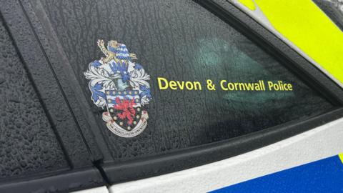 A close up shot of the window of a Devon & Cornwall Police car on a rainy day. The force's logo is visible as well as water dripping down the side of the car.