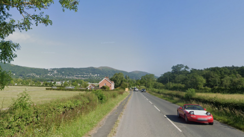 The straight road stretches out into the countryside with hills in the background. There is a house on the left hand side and cars driving on either side.
