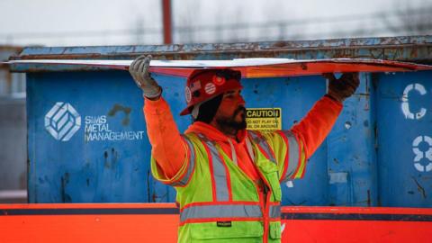A bearded construction worker wearing an orange sweatshirt and yellow safety vest carries a sheet of construction material over his his head. In the background there is a a blue container. The picture was taken at the site of the Gateway Tunnel construction project as seen from North Bergen, New Jersey, on February 10, 2026.
