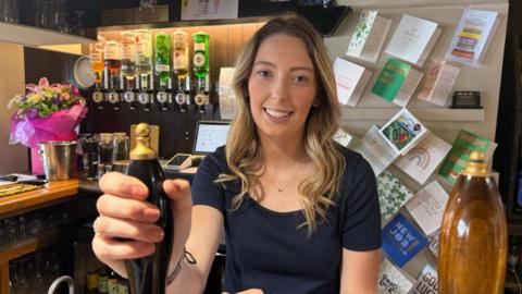 A woman, Neve, stands behind the bar in a pub. There are bottles of spirits and good luck cards behind her