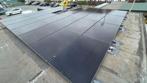 A view of black solar panels on top of a building. The roof underneath is grey. On the ground level to the left there is a car park with parked cars.
