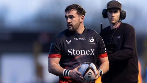 James Hadfield holds a rugby ball in both hands in front of him, with a cameraman dressed all in black in the background