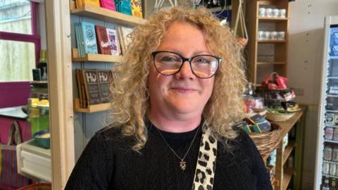 Cherylee Barker smiles at the camera. She is stood in a cafe which shelves and chocolate bars in the background,