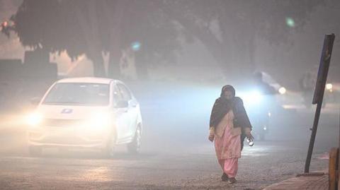 A woman wearing a pink traditional Indian dress and a black scarf walks along the side of the road in the early hours of the morning in Karol Bagh, Delhi, as vehicles pass by with their lights on due to low visibility.