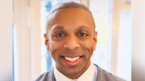 Martyn Latchman: a professional head‑and‑shoulders portrait of a man indoors. He is wearing a light shirt under a grey jacket, and looking directly at the camera with a smiling expression.