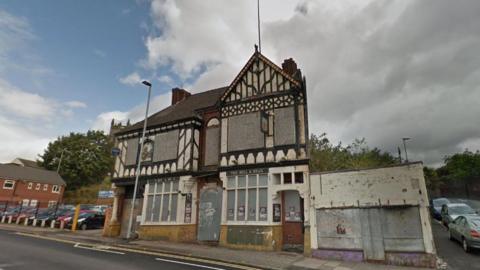 A street view image of an Edwardian pub building, which is empty and derelict. The windows and doors are boarded up. A car park is visible on the left. Cars can also be seen parked along the street on the right.