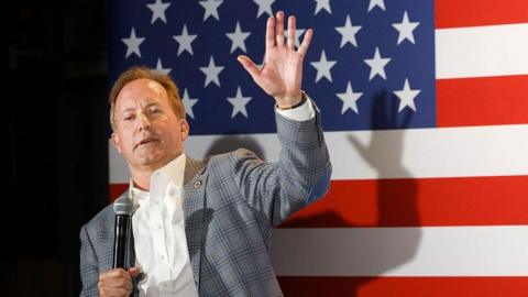 Texas Attorney General Ken Paxton waves while leaning and holding a microphone in front a US flag backdrop.