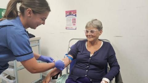 A woman in a blue top sits in a chair while a medic in blue scrubs takes her blood