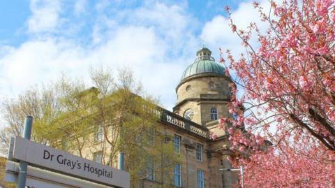 Hospital building with a 'Dr Gray's' sign and some pink blooms on a tree to the right of the image.