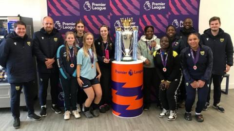 A team of young female football players poses with football coaches next to the Premier League trophy