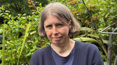 Sharon Morgan, a woman aged 50 standing in the front garden of her house. She has honey blonde bobbed hair and is wearing a navy top and navy cardigan.