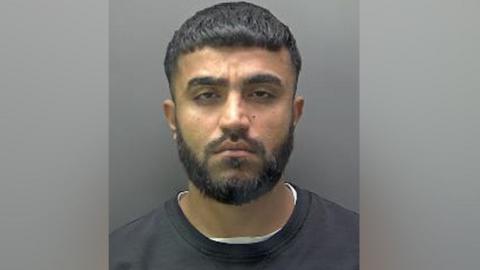 A mugshot of Galmne, wearing a white t-shirt under a black sweater. He has a black beard and moustache and short dark hair. 