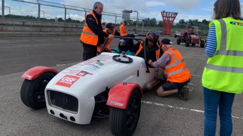 Racing car on track with two people seated and others standing next to it