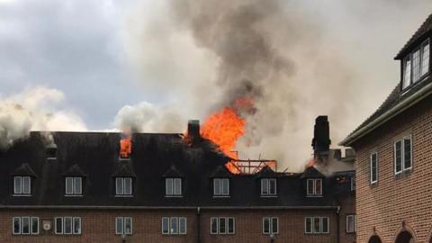 Orange flames and plumes of black smoke rise from a hole in the roof of the hall. The long brown brick building is two storeys tall.