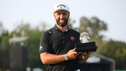 Jon Rahm, with a dark beard and wearing a black polo shirt, smiles as he holds the LIV Mexico trophy - a black base with a glass skull
