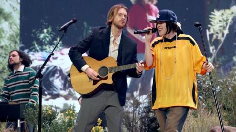 Billie Eilish performs at the Grammy Awards alongside her brother Finneas, with Finneas playing an acoustic guitar. Billie is wearing a yellow sports top, a long necklace and a blue baseball cap. Finneas has a blue jacket, white shirt and grey trousers. A keyboardist can be seen in the background. 