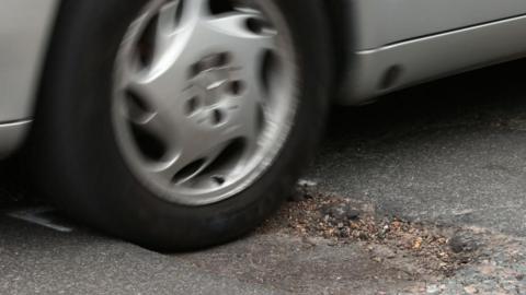 A close up of car wheel hitting a pothole in the road. The car is silver. 