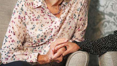 An older woman wearing a floral shirt and sitting in a chair holds hands with someone wearing a black and white dotted shirt.