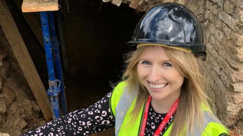 A woman in a hard hat and a high-vis yellow vest proudly squatting in front of an opening into a tunnel. She is smiling.