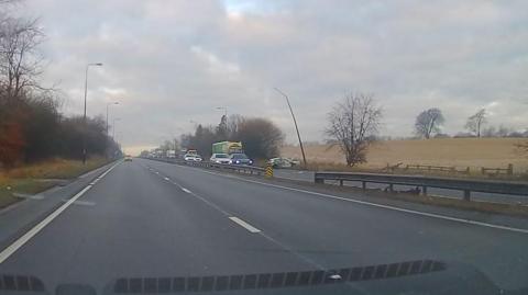 View from a car windscreen of a road stretching ahead. On the right hand - opposite - carriageway there is stationary traffic ahead behind what appears to be a damaged police car just off the road