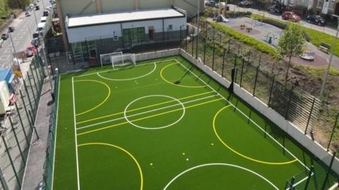 An artists impression of the playzone showing a bright green playing field surrounded by high fences. 
