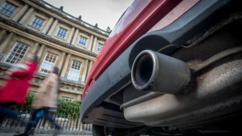 A red car's exhaust pipe is focused on while two people in the background walk past.