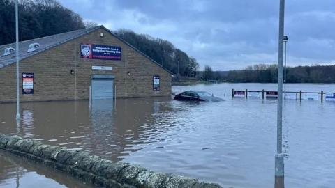 A brick clubhouse building on the left half of the screen, about 15 metres away from the camera. It has flooded to approximately knee-height. A car is to the right of the building and has flooded up to the bonnet. The rest of the image shows flooding. A brick wall near the camera just about pokes above the water. There area trees in the distance.