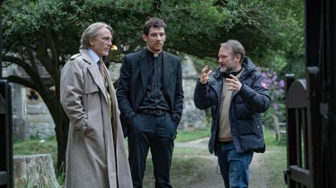 (Left to right) Daniel Craig and Josh O'Connor and writer-director Rian Johnson are standing just beyond the entrance to a church graveyard with the church itself in the background. Johnson, on the right, is gesturing to Craig and O'Connor, who are listening intently. Craig is wearing a long beige raincoat with shirt and tie, while O'Connor is dressed as a clergyman in dog collar and black shirt, jacket and trousers. 