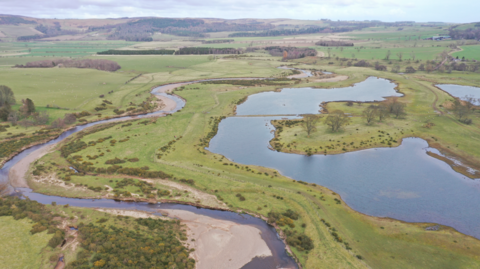 An aerial view of a small river which meanders its way through fields with a large pond area to the right-hand side 