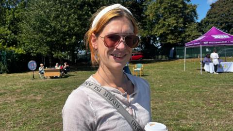 The image features a woman with ginger hair, wearing a headband and sunglasses, holding a cup of coffee while standing in a field. In the background, green trees frame several gazebos hosting various organisations.
