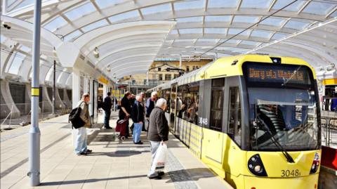 Passengers wait at a platform to get on a Bee Network yellow coloured tram.