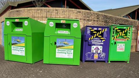 Image of four clothes and shoes recycling banks, lined up against a brick wall. In the background can be seen the roof of a building.