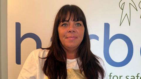 Danielle Chadwick is a woman with long dark brown hair and a straight fringe. She is wearing a white and gold t-shift and is smiling at the camera. Part of the Harbour logo on the wall behind her.
