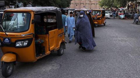 A woman covered with a long robe from head to toe, with her face covered by a medical mask, walks down the street near a tuktuk. Men can be seen in the background