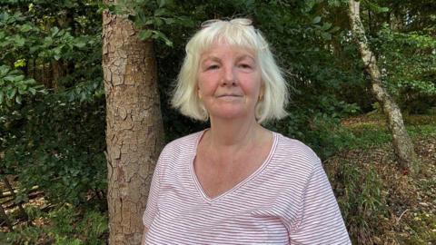 A woman with sort white blonde hair, and glasses on her head. She is wearing a pink and white striped top and is standing in an area of woodland