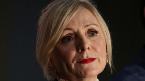 A close up shot of a blonde haired woman, aged in her 50s or so, as she looks into the distance.