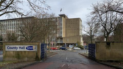 Entrance gates of Durham County Council with a sign reading 'County Hall' and the council building in the background