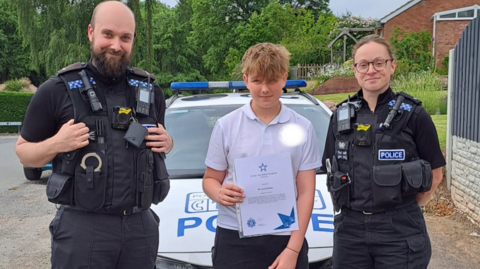 Joey stands in between two police officers in uniform. They are stood in front of a police car by some houses with Joey holding a certificate. He has short brown hair and is wearing a polo top and trousers.