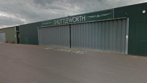 The outside of an aircraft hangar at Shuttleworth in Biggleswade. The outside of the building is dark green and there is a sign on top which reads SHUTTLEWORTH in white lettering.