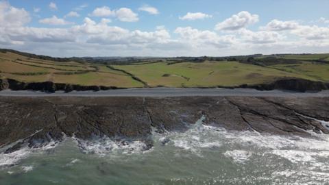 A aerial view of the coastline, showing waves breaking onto a rocky foreshore. there are gently sloping fields beyond