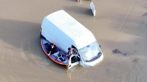 A white van stuck in brown water which is up to its headlights. A person is leaving the van and getting into a canoe.