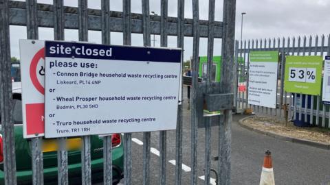 White and navy blue sign on the entrance to the St Austell Household Waste and Recycling Centre site says "site closed" and tells people to use three alternative locations. A green Mini Cooper is parked the other side of the main gate.