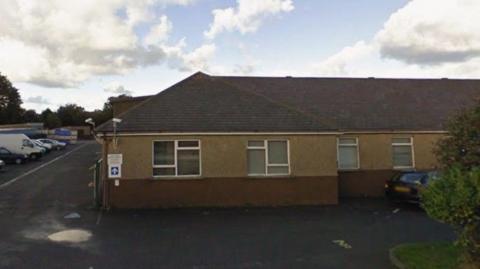 A single storey brown building with white rimmed windows. There are cars parked to the left of it.