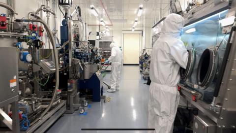 The inside of a factory with large silver machinery. There are two people in white PPE protective wear and wearing blue gloves and blue masks.