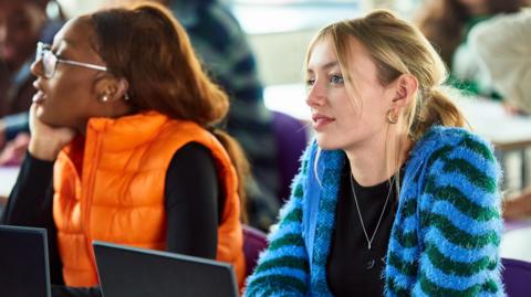 Two women sittign in front of laptops. One is wearing an orange gilet - she is looking off to the side. The main woman in view is wearing a green and blue stripey cardigan, has blond hair pulled back in a ponytail.