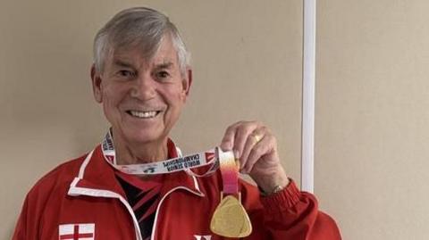A man smiling holding his medal. He is wearing a red jacket. 